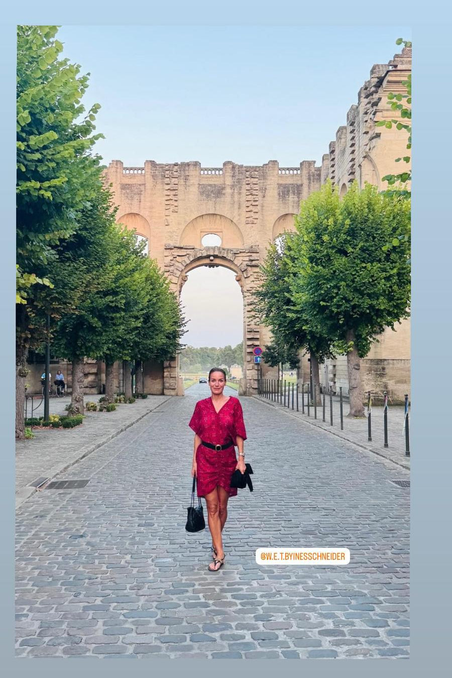Woman in a red dress walking through an archway with trees on either side.